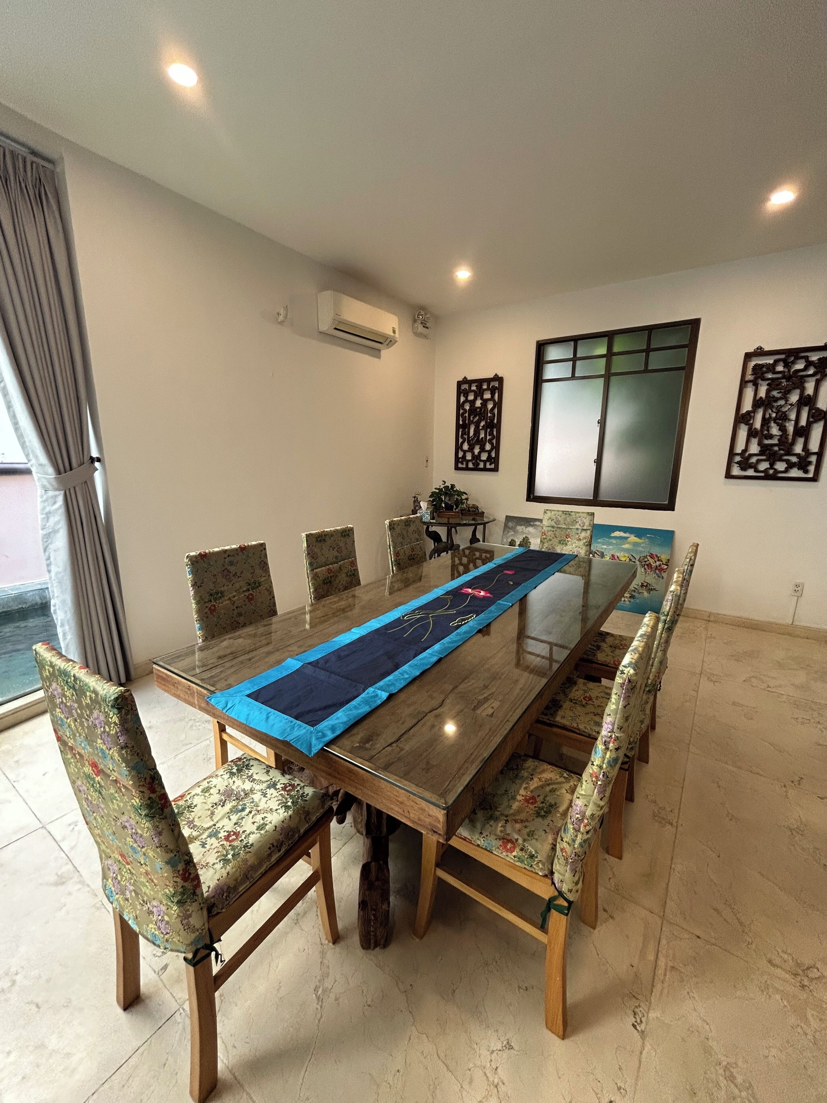 Uma área de refeições com uma mesa comprida, cadeiras florais, um corredor decorativo e uma janela com vista.
