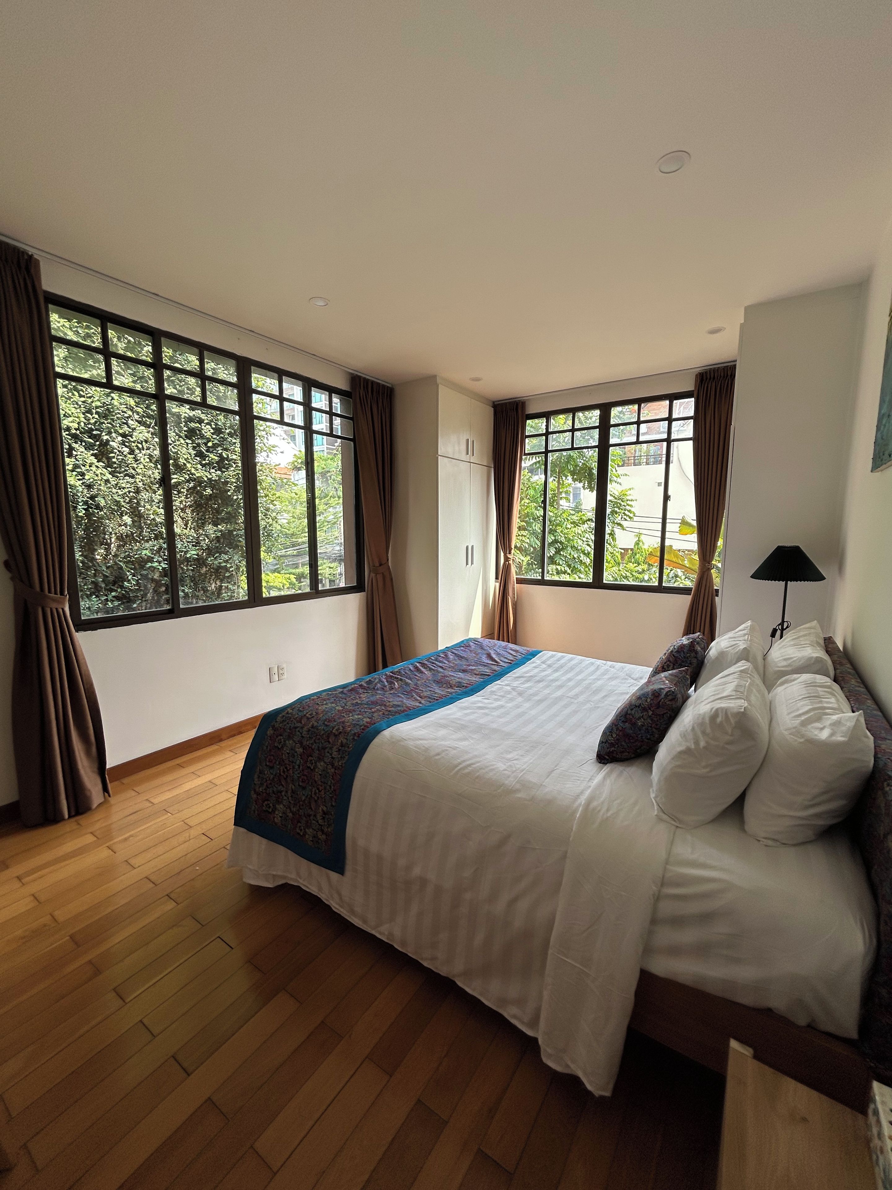 A bedroom with a large bed, a nightstand, a lamp, and a view of greenery through the windows.