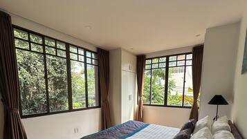 A bedroom with a large bed, a nightstand, a lamp, and a view of greenery through the windows.