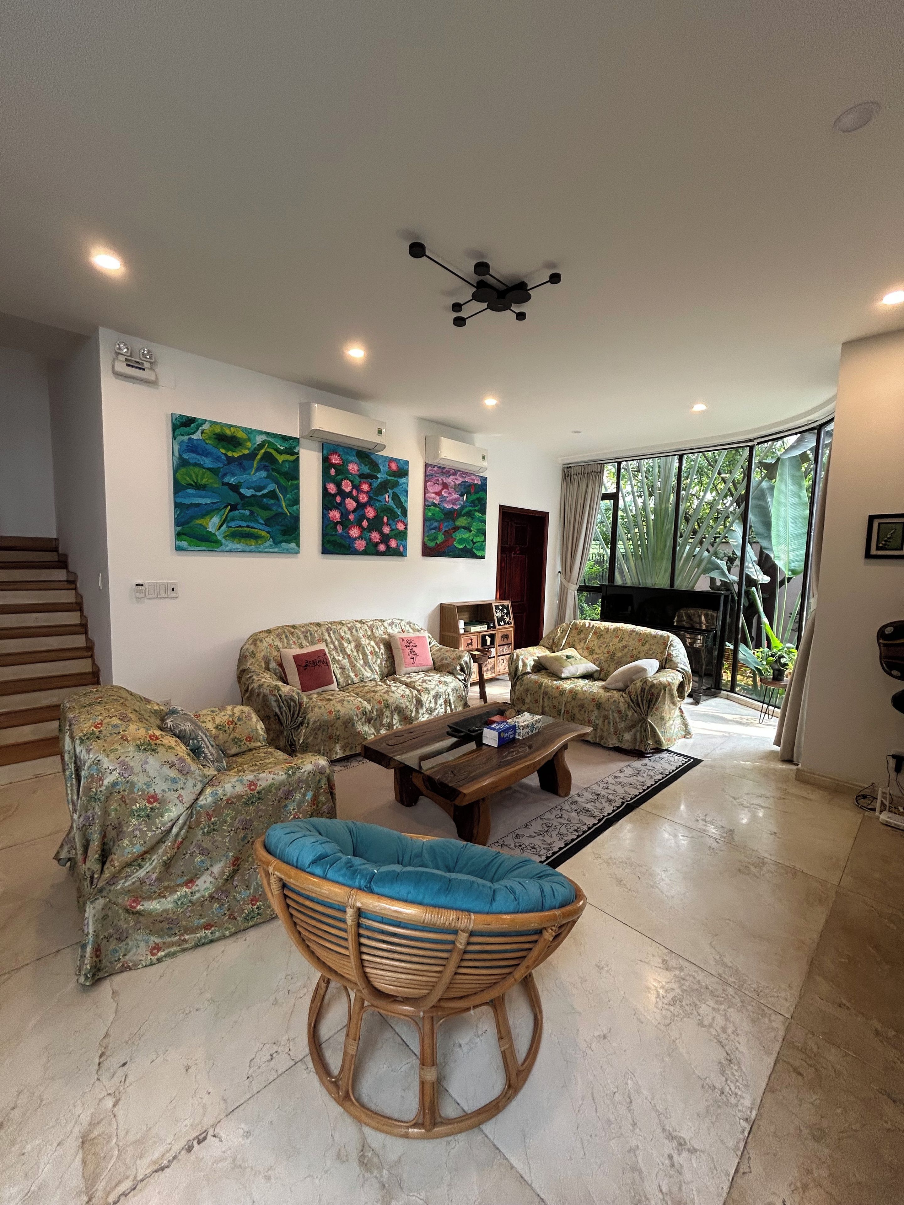 A living room with a sofa set, a coffee table, and a wicker chair. There are paintings on the wall and a ceiling fan.