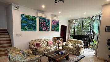 A living room with a sofa set, a coffee table, and a wicker chair. There are paintings on the wall and a ceiling fan.