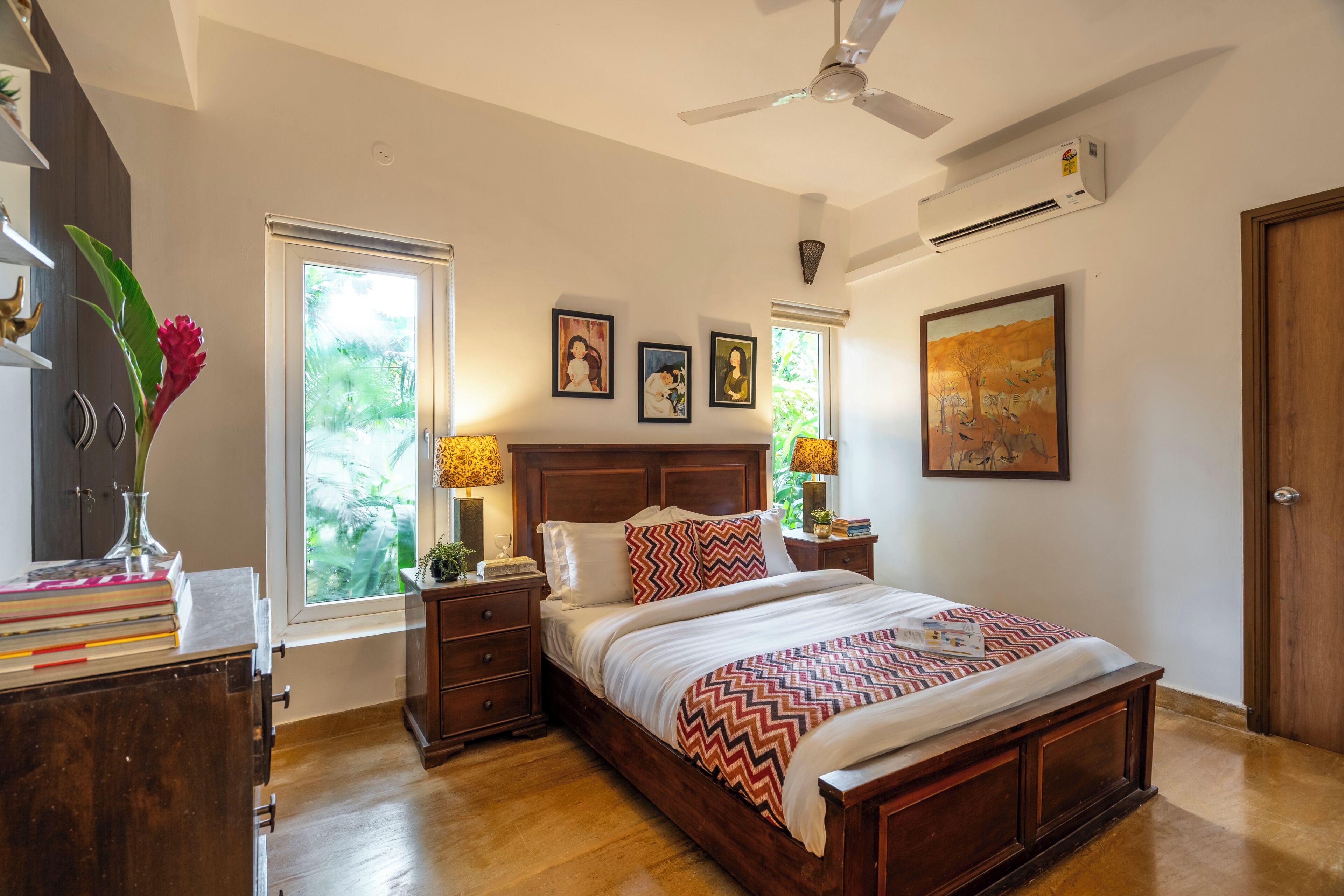A bedroom with a wooden bed, bedside tables, a nightstand, a ceiling fan, and a window with a view of greenery.