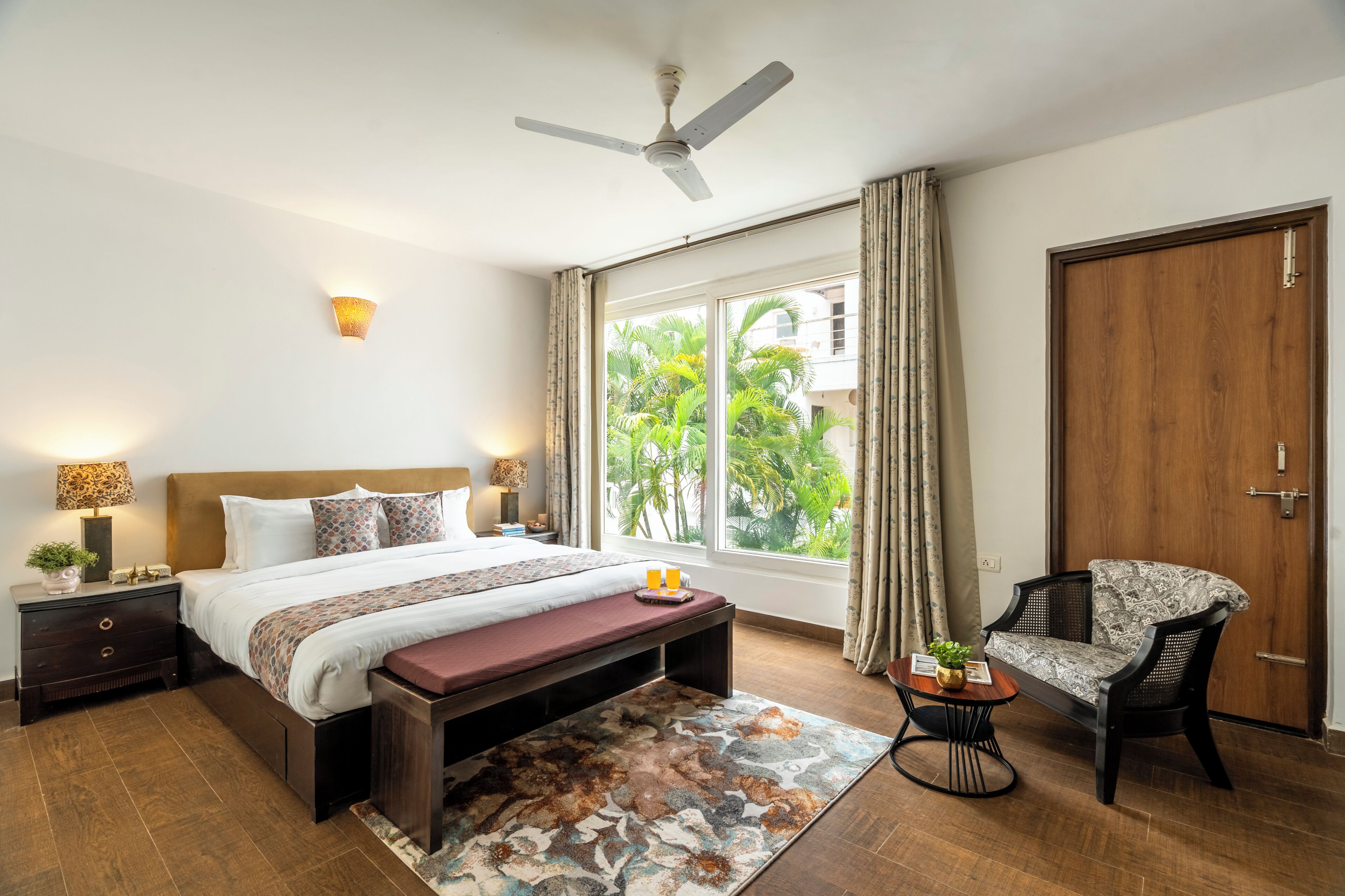 Um quarto com cama, mesas de cabeceira, um banquinho, uma cadeira, ventilador de teto, janela com cortinas e vista para o verde lá fora.