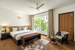 A bedroom with a bed, bedside tables, a bench, a chair, a ceiling fan, a window with curtains, and a view of greenery outside. - Staymaster Taram - 4BR Villa in Candolim (Candolim)