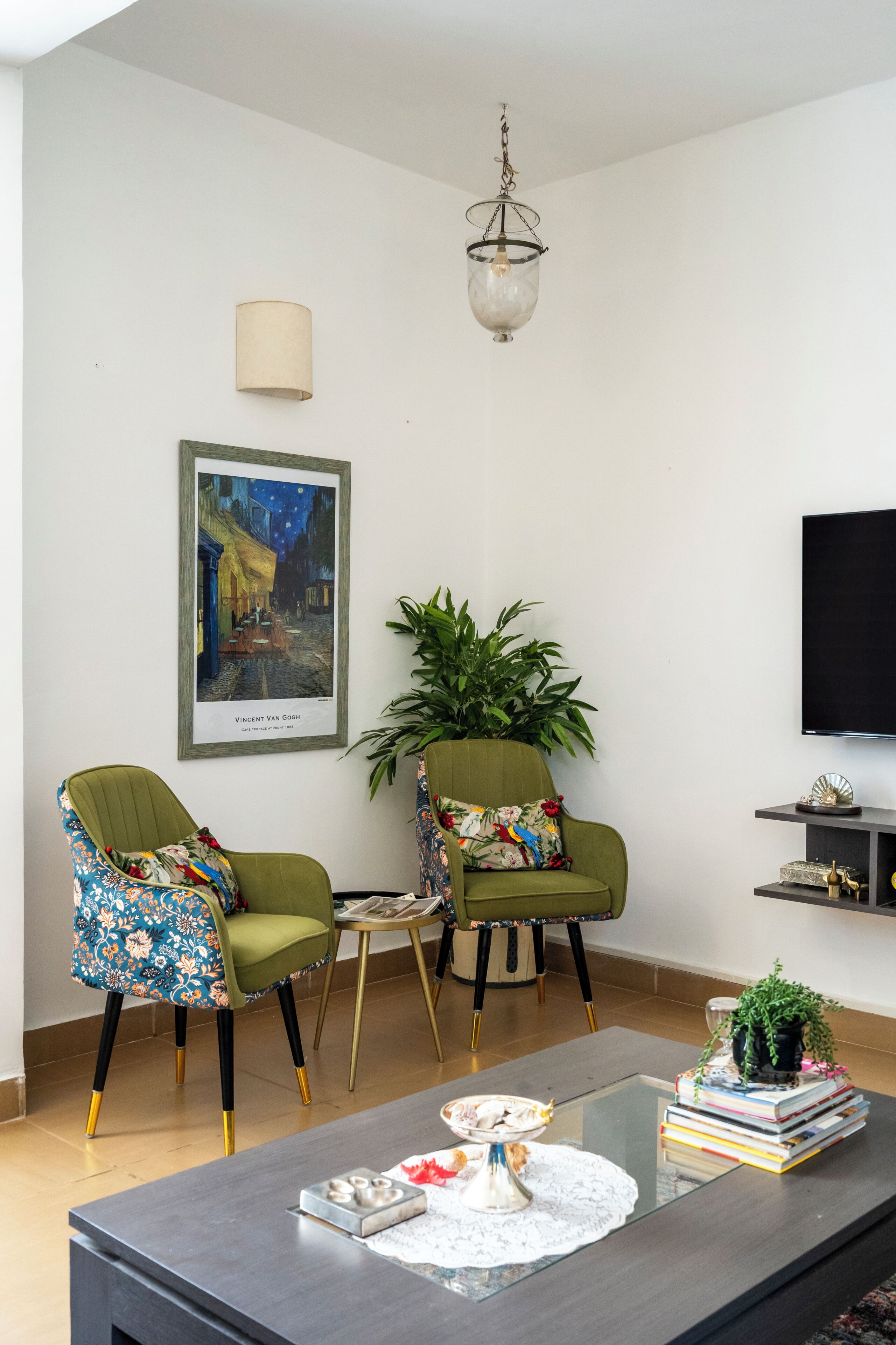 Uma sala de estar com dois poltronas verdes, uma mesa de centro, um quadro emoldurado, uma planta em vaso e uma televisão na parede.