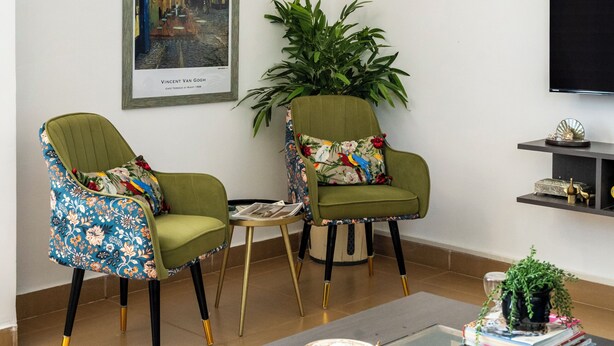 A living room with two green armchairs, a coffee table, a framed painting, a potted plant, and a television on the wall.