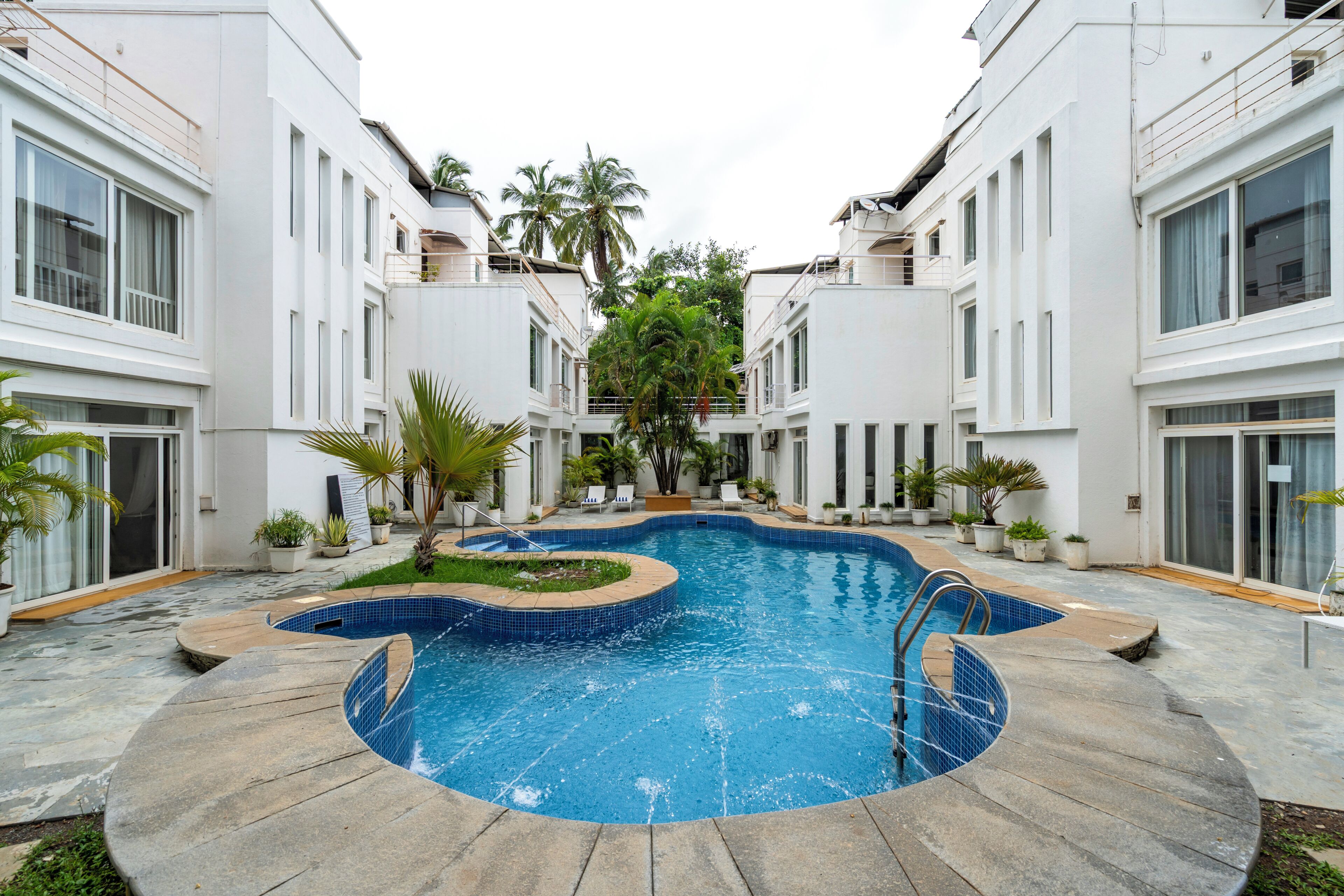 A swimming pool surrounded by modern buildings with balconies, potted plants, and a clear sky.