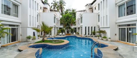 A swimming pool surrounded by modern buildings with balconies, potted plants, and a clear sky.
