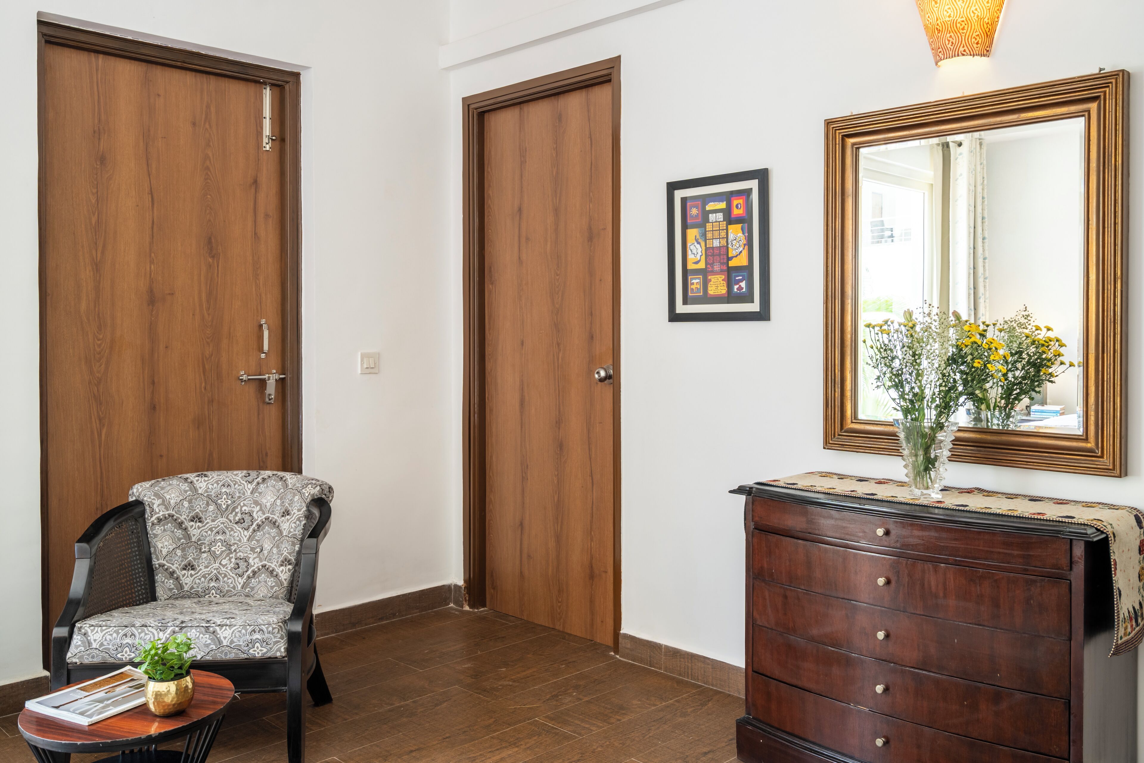 A hallway with two wooden doors, a chair with a patterned cushion, a small table with a plant, a dresser with a mirror, and a framed picture on the wall.
