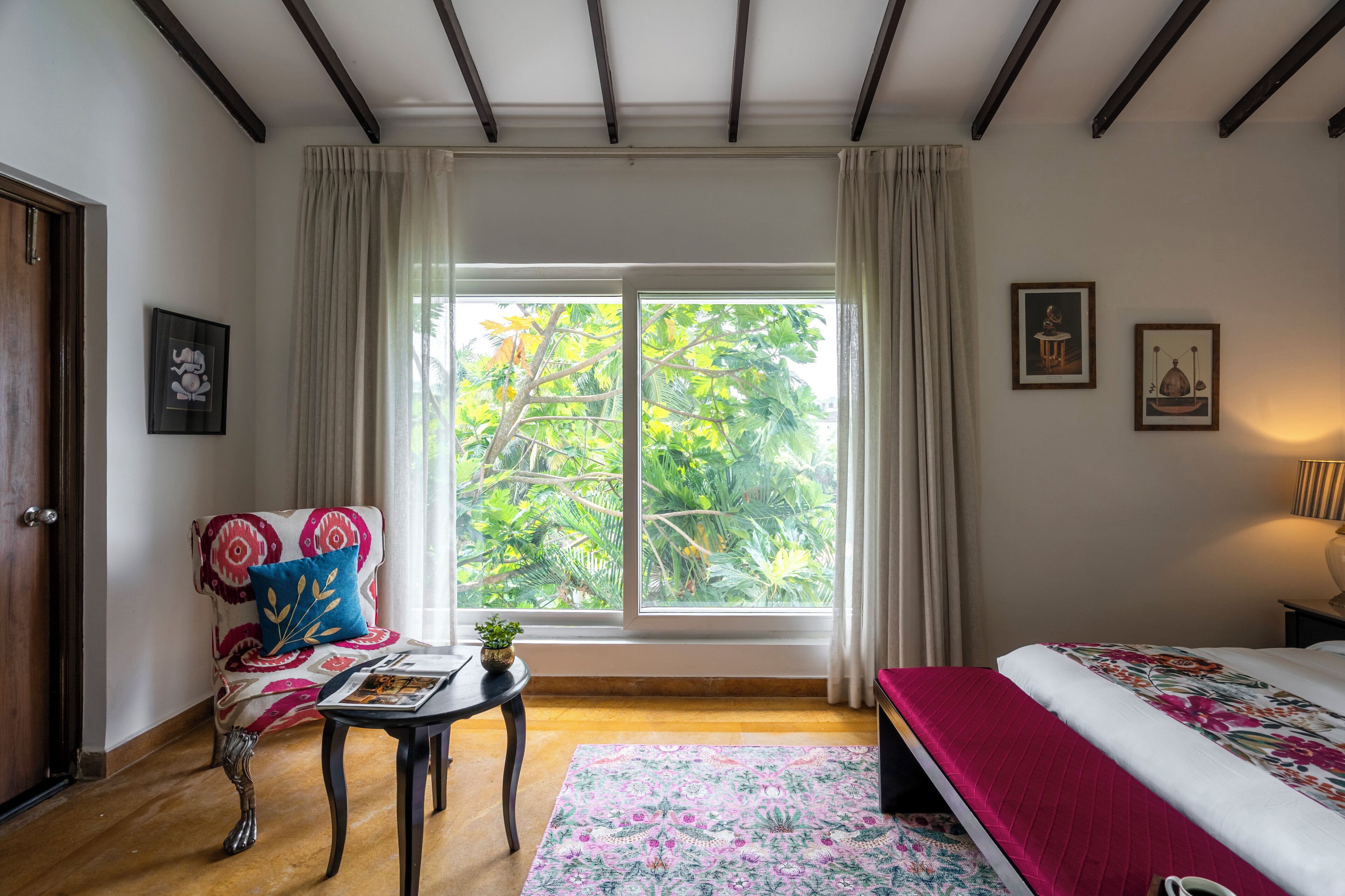 Ein Schlafzimmer mit großem Fenster, einem Bett, einem Sessel mit gemustertem Kissen, einem kleinen Tisch mit Pflanze und einem gerahmten Bild an der Wand.
