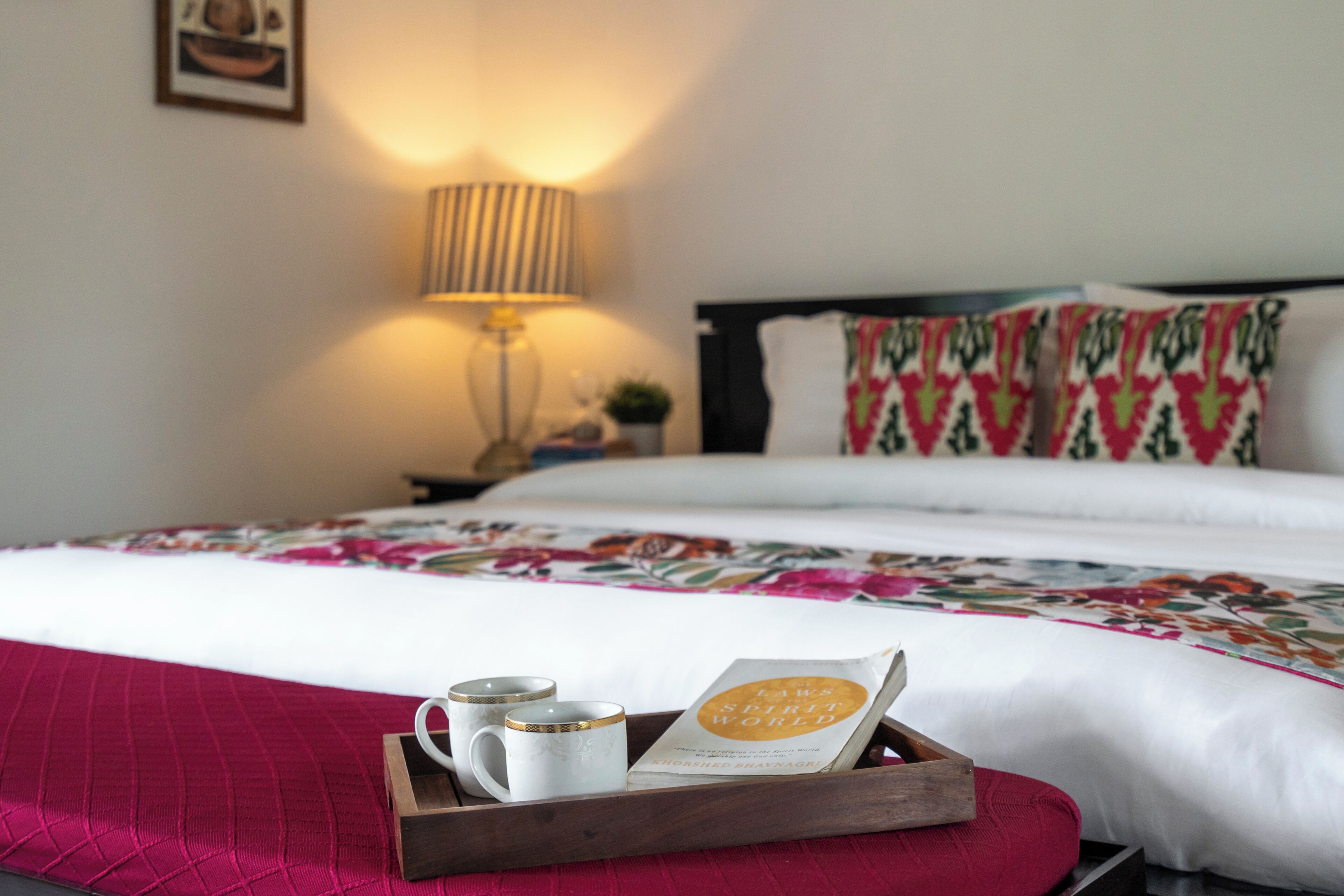 Una cama bien hecha con una colcha floral, una lámpara de noche y una bandeja con un libro y dos tazas.