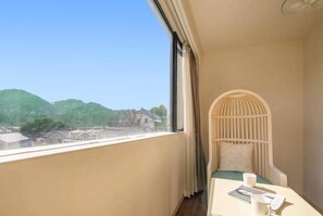 A balcony with a view of a green landscape and buildings, a cushioned seating area, and a table with a book and a mug.