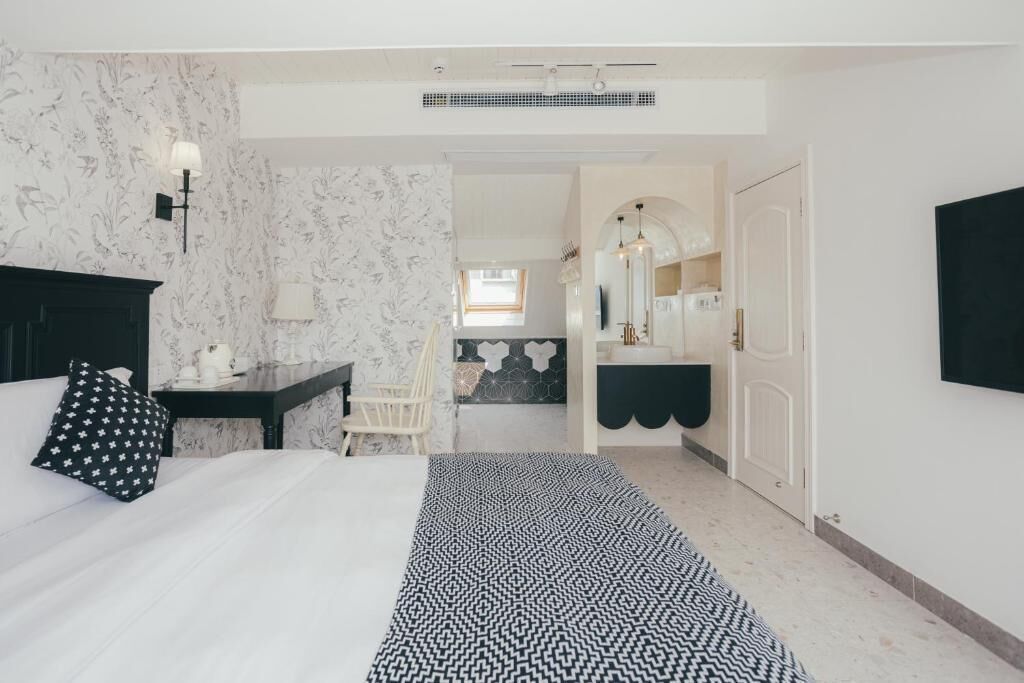 A hotel room with a bed, a desk, a chair, a bathroom with a mirror and sink, and a hallway leading to another room.