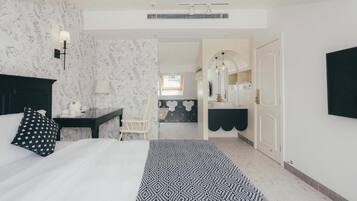 A hotel room with a bed, a desk, a chair, a bathroom with a mirror and sink, and a hallway leading to another room.