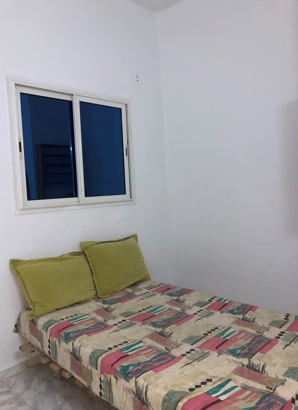 Una habitación pequeña con una cama individual, pared blanca lisa, ventana con persianas bajadas y una colcha de colores.