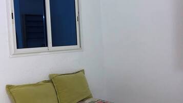Una habitación pequeña con una cama individual, pared blanca lisa, ventana con persianas bajadas y una colcha de colores.