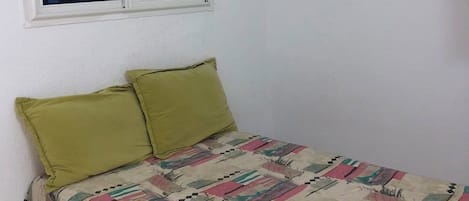A small, single-bed room with a plain white wall, a window with closed blinds, and a colorful bedspread.