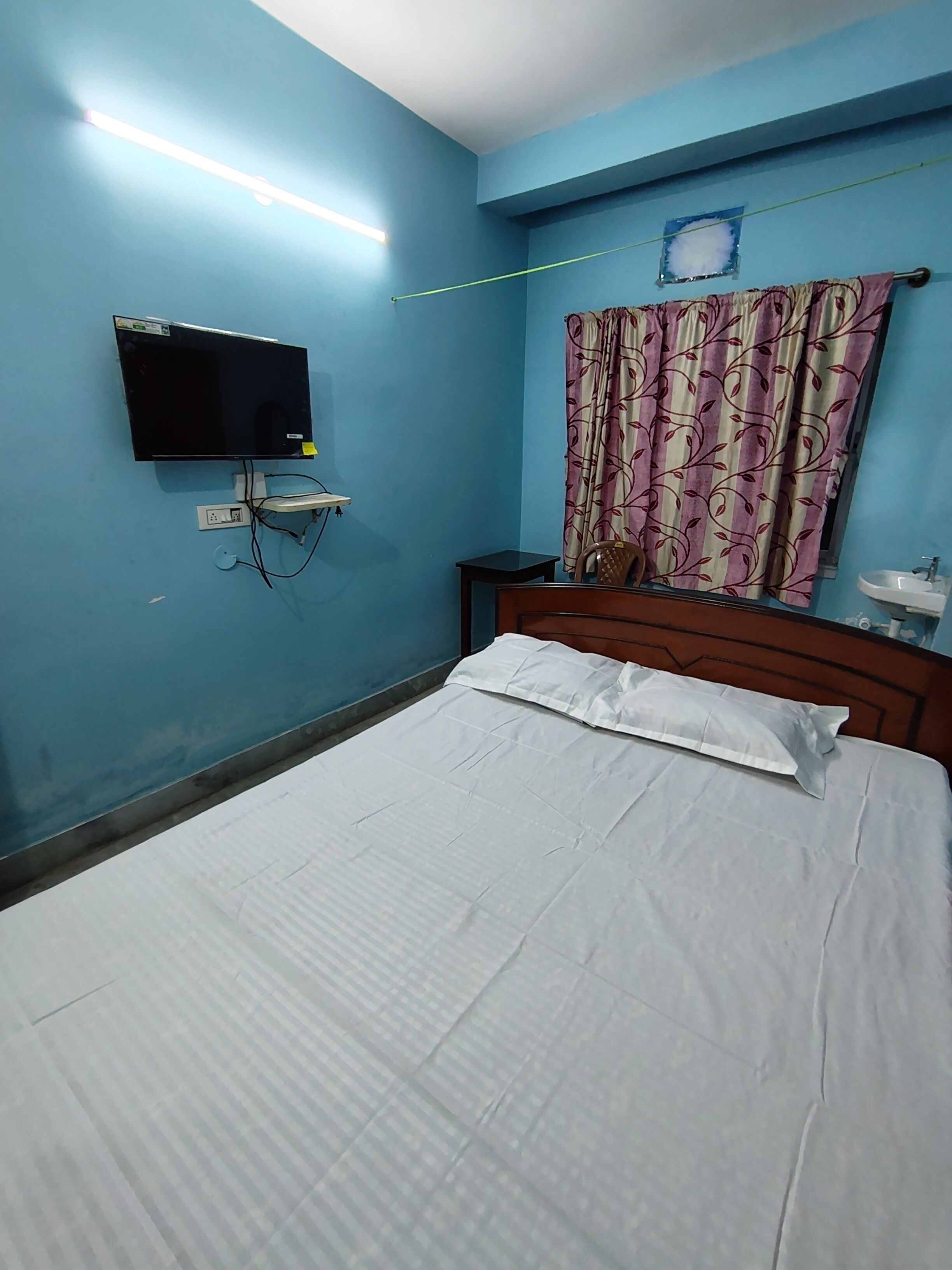 Un dormitorio con una cama, televisor montado en la pared, una mesita de noche, una silla y una cortina.