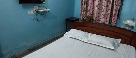 Un dormitorio con una cama, televisor montado en la pared, una mesita de noche, una silla y una cortina.