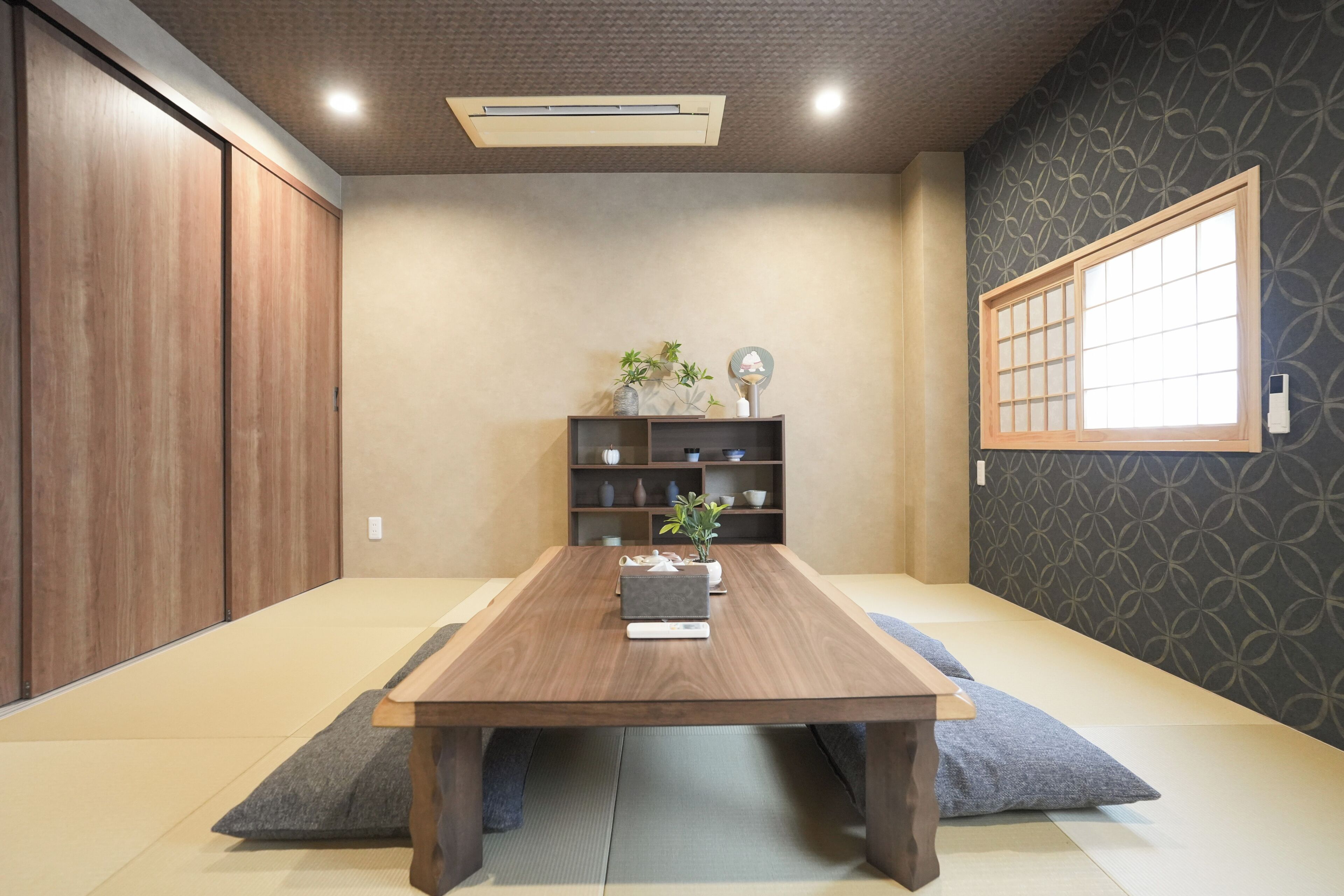 A room with a wooden table, a shelf with decorative items, a window with a grid pattern, and a patterned wall.