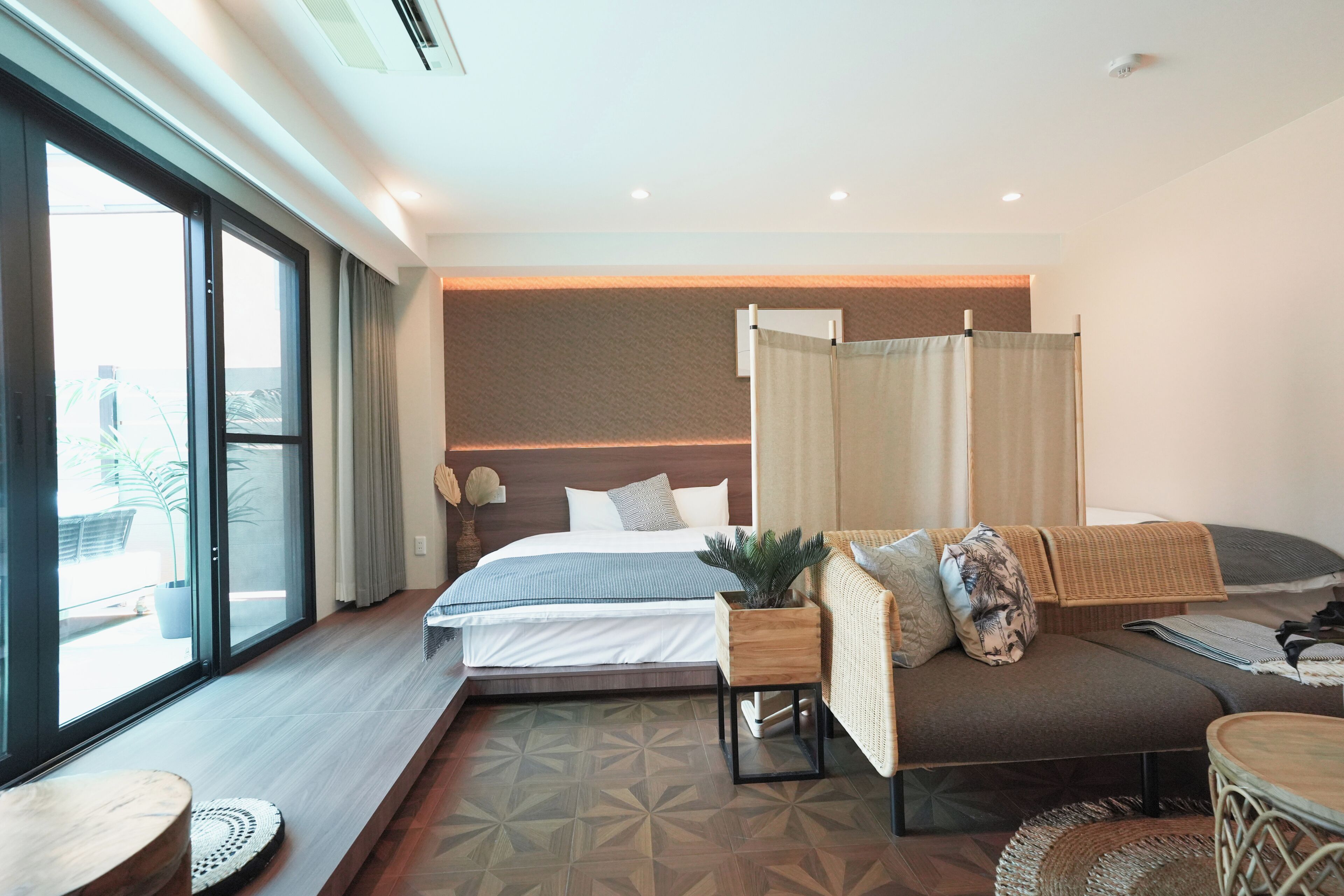 A modern hotel room with a large bed, a sofa, a small table, and a view through a large window.