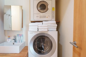 A front view of a modern laundry room with a stacked washer and dryer, a sink, and a mirror. - SERENE | Kuromon ・ 201 | 4BR ・ 2 Shower Rooms | Private Home for Families and Groups (Osaka)