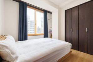 Ein Schlafzimmer mit Bett, Kleiderschrank, einem Fenster mit Vorhängen und Blick auf Gebäude.