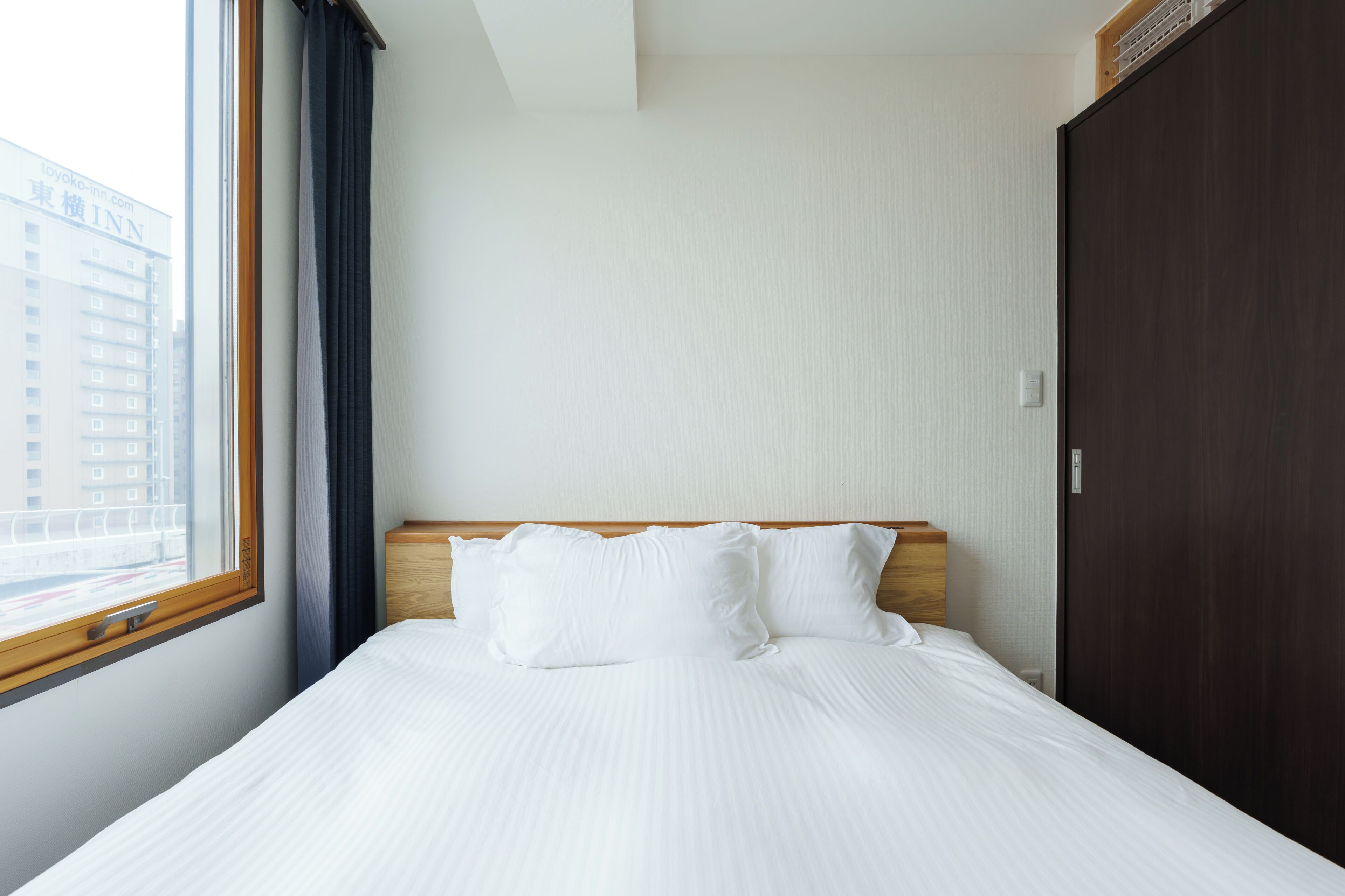 A hotel room with a bed, a wooden headboard, a window with curtains, and a view of a building outside.