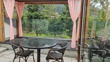 Un patio cubierto con una mesa redonda y sillas, barandilla de vidrio y vista a la vegetación.