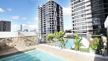 A rooftop swimming pool with a stone wall and palm trees, surrounded by high-rise buildings.