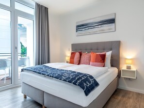 A modern bedroom with a large bed, a grey headboard, and a view of the outdoors through a sliding glass door. - Parkidylle mit Sauna (Lübeck)