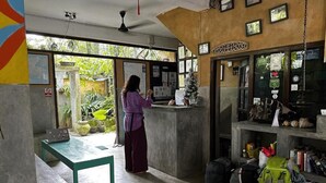 Zona de recepción con mostrador, una mesa verde y una mujer de pie. Hay bolsas y una mochila verde en el suelo.