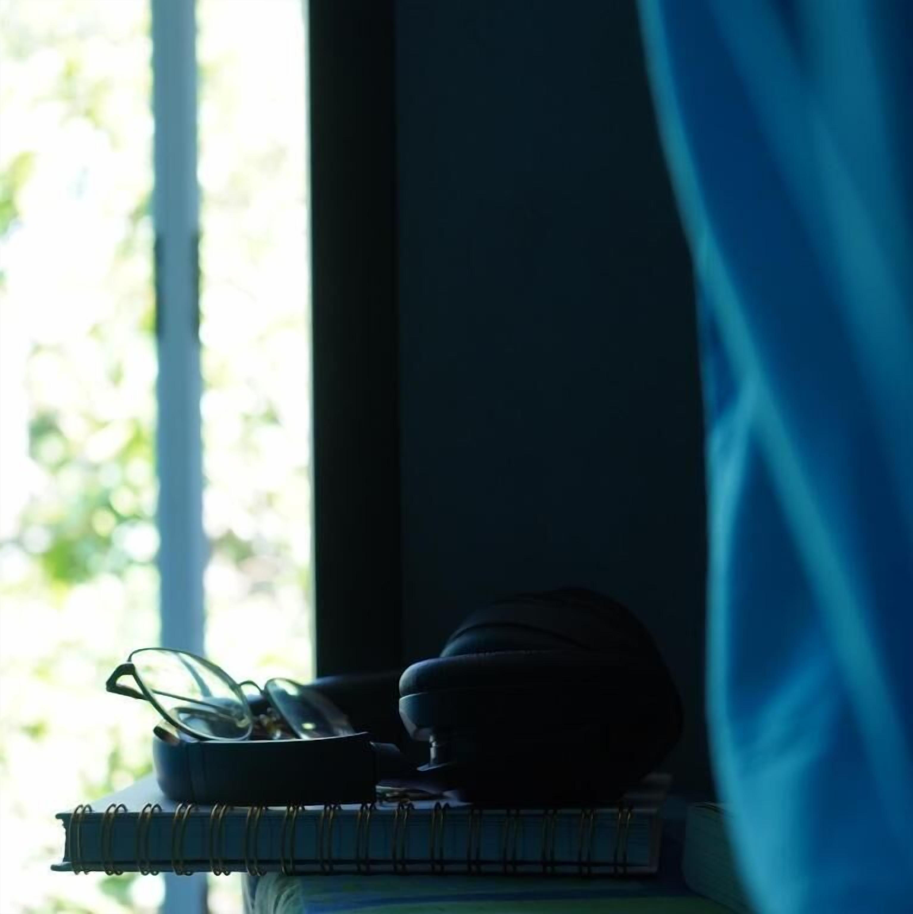 A window with a view of greenery, a notebook, and a pair of glasses on a surface.