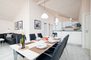 A modern dining area with a long table set for six, a sofa, and a kitchen in the background.