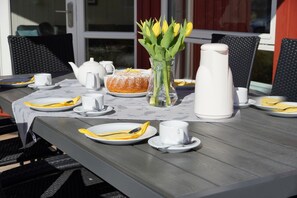 Ein Tisch im Freien, gedeckt mit Tellern, Besteck, einer Teekanne, Zuckerdose, einer Karaffe und einer Vase mit Blumen.