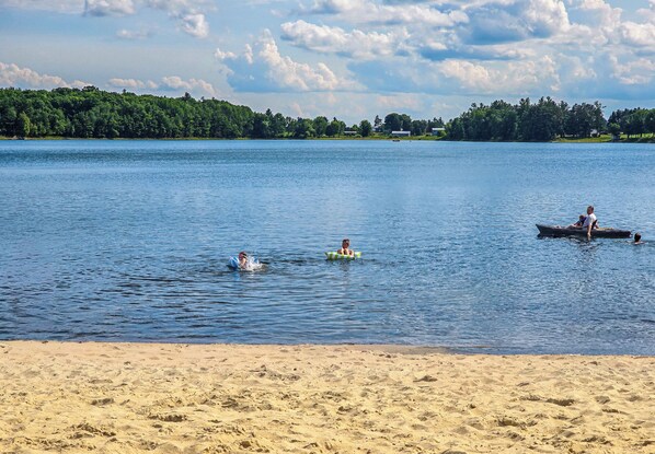 A beach with calm water, a sandy shore, and a distant tree line. - Windfall Lake | Sandy Beach | Hot Tub | Game Room | Kayaks | Ski & Snowmobile (Exeland)