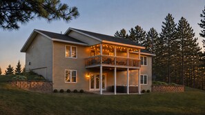 A two-story house with a balcony, a stone retaining wall, and a grassy hill. - Windfall Lake | Sandy Beach | Hot Tub | Game Room | Kayaks | Ski & Snowmobile (Exeland)