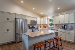 A modern kitchen with stainless steel appliances, white cabinetry, a large island with bar stools, and a view of greenery outside.