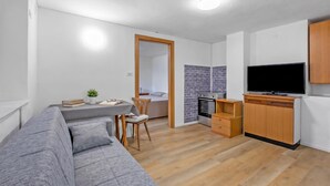 A modern living room with a grey sofa, a dining table with chairs, a TV, and a kitchen area with a stove and oven. - Falesia Vertical Rock View (Arco)