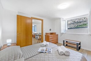 Ein Schlafzimmer mit einem Bett, einem Nachttisch mit Lampe, einem Schminktisch, einer Bank und einem Fenster mit Blick auf eine gebirgige Landschaft.