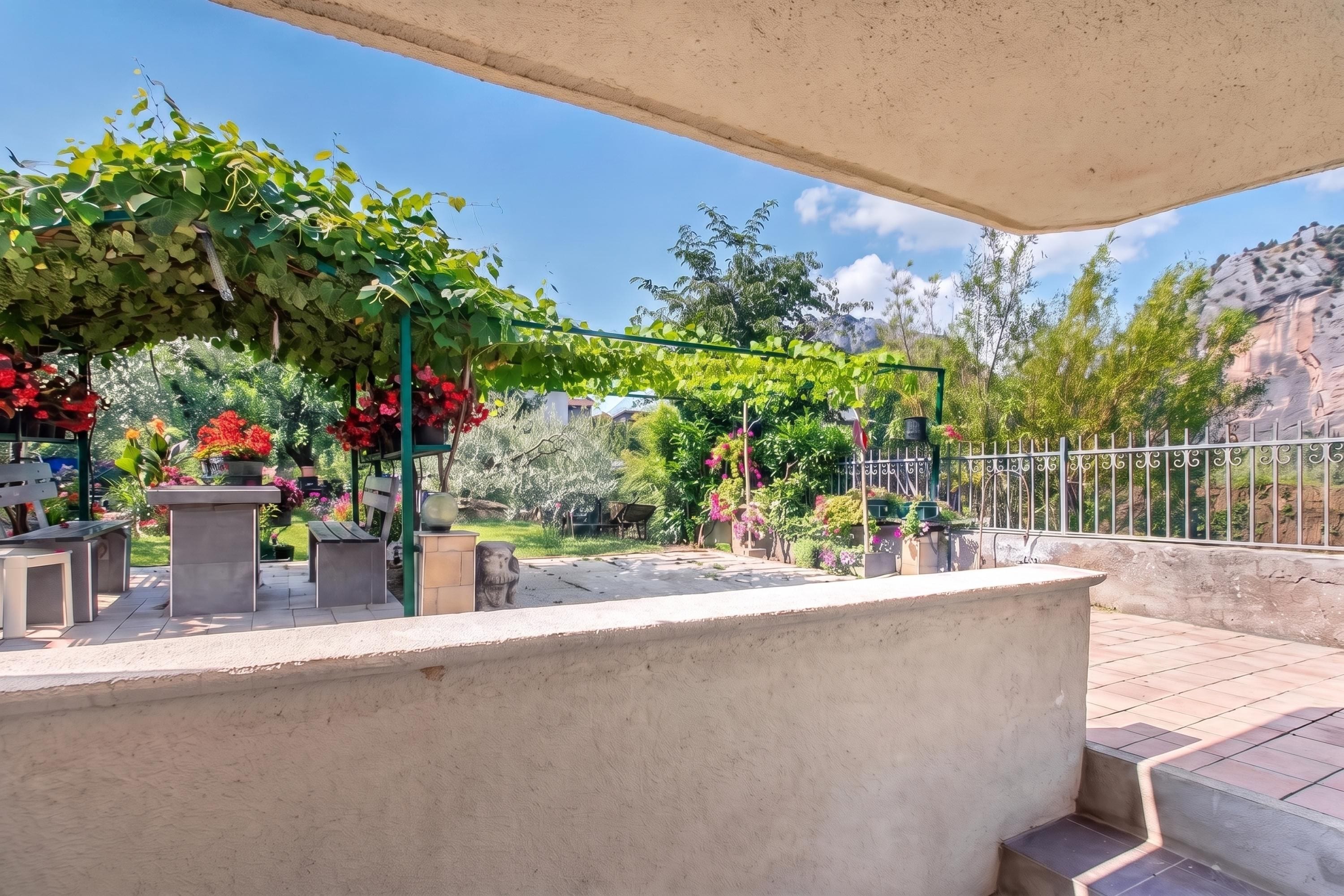 Een terras buiten met een pergola begroeid met groene bladeren, zitgedeeltes en uitzicht op een bergachtig landschap.
