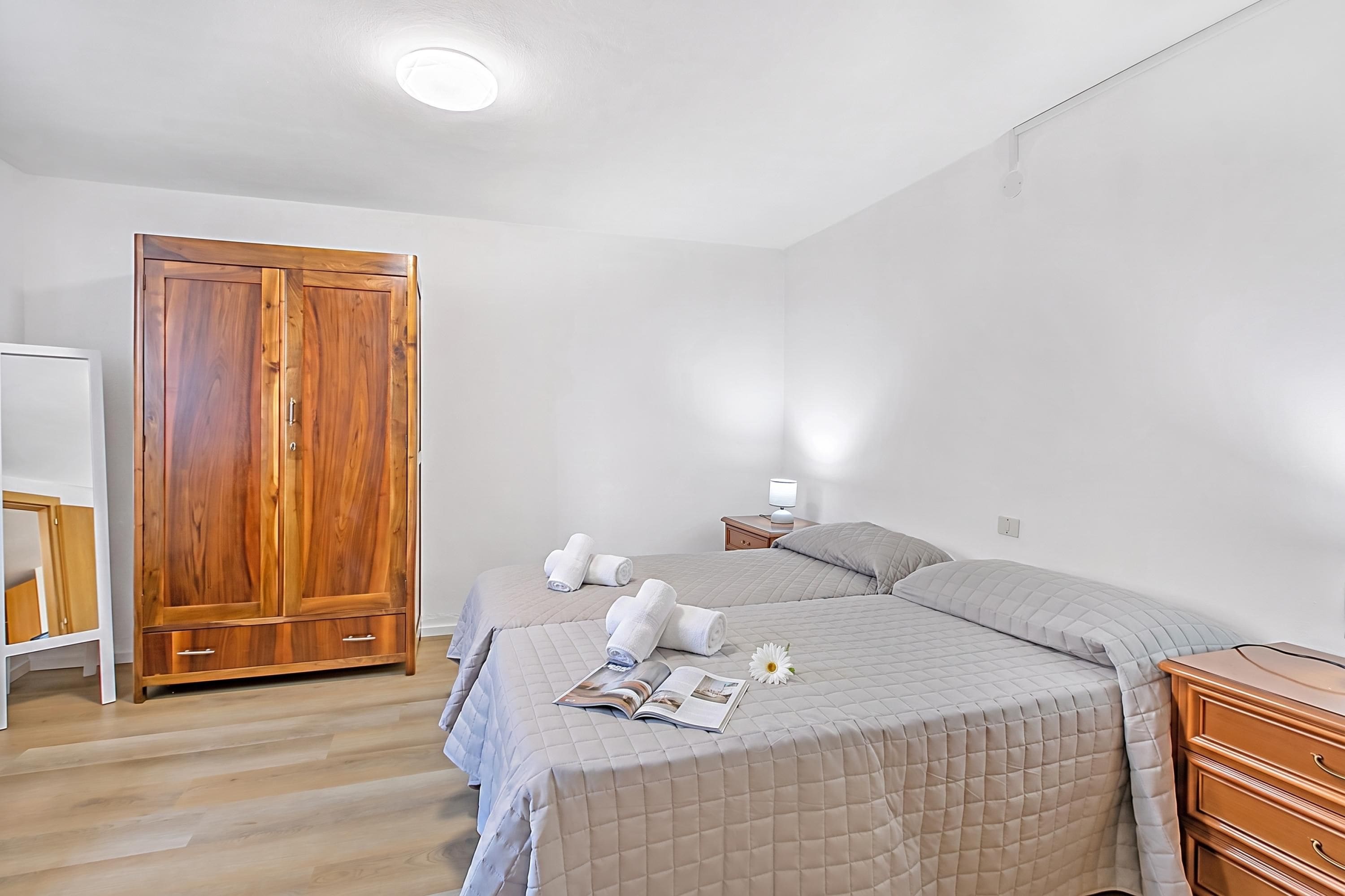 A bedroom with a double bed, a wooden wardrobe, a bedside table with a lamp, and a small nightstand.