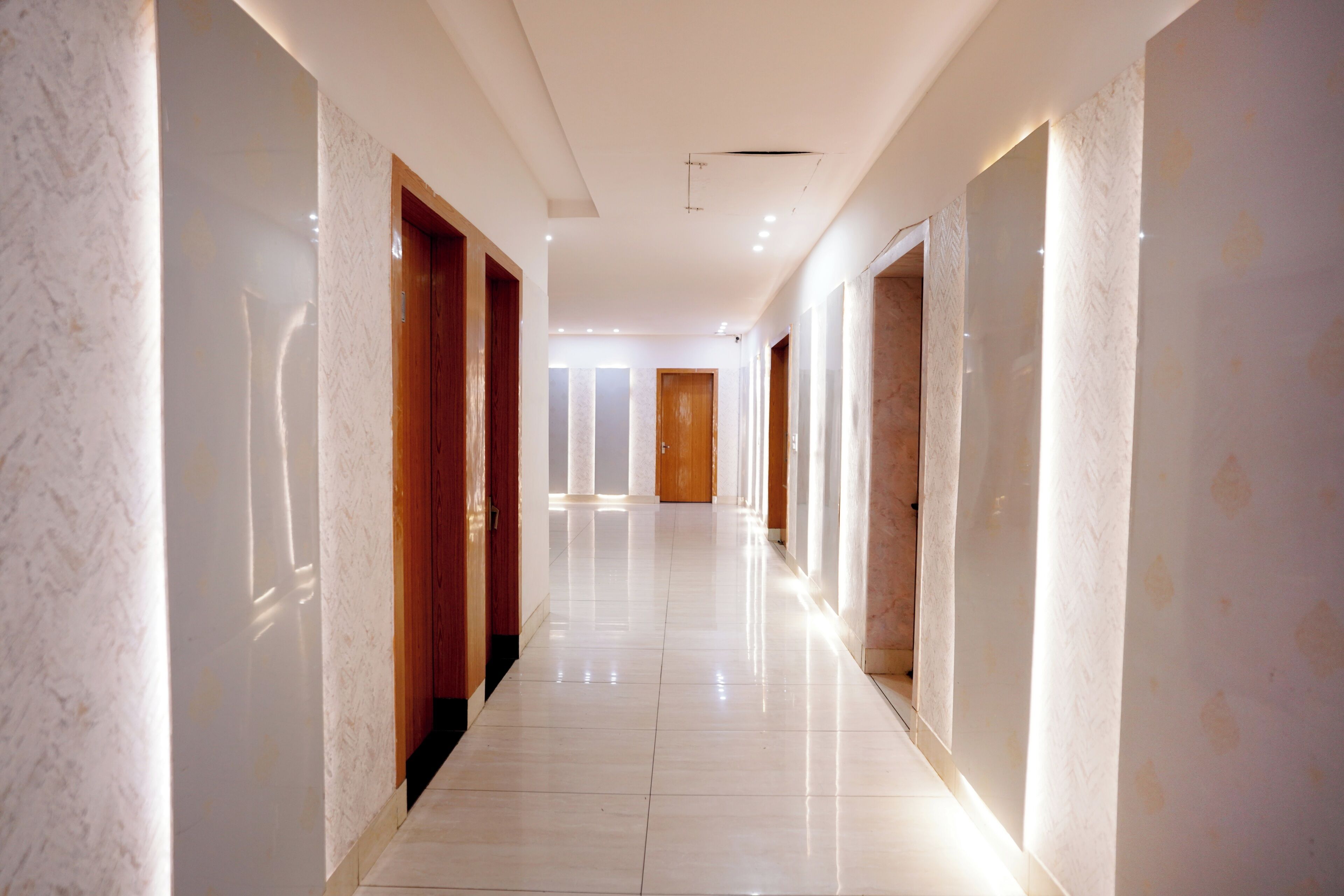 A long, well-lit corridor with marble walls and wooden doors, leading to a room with a large window.