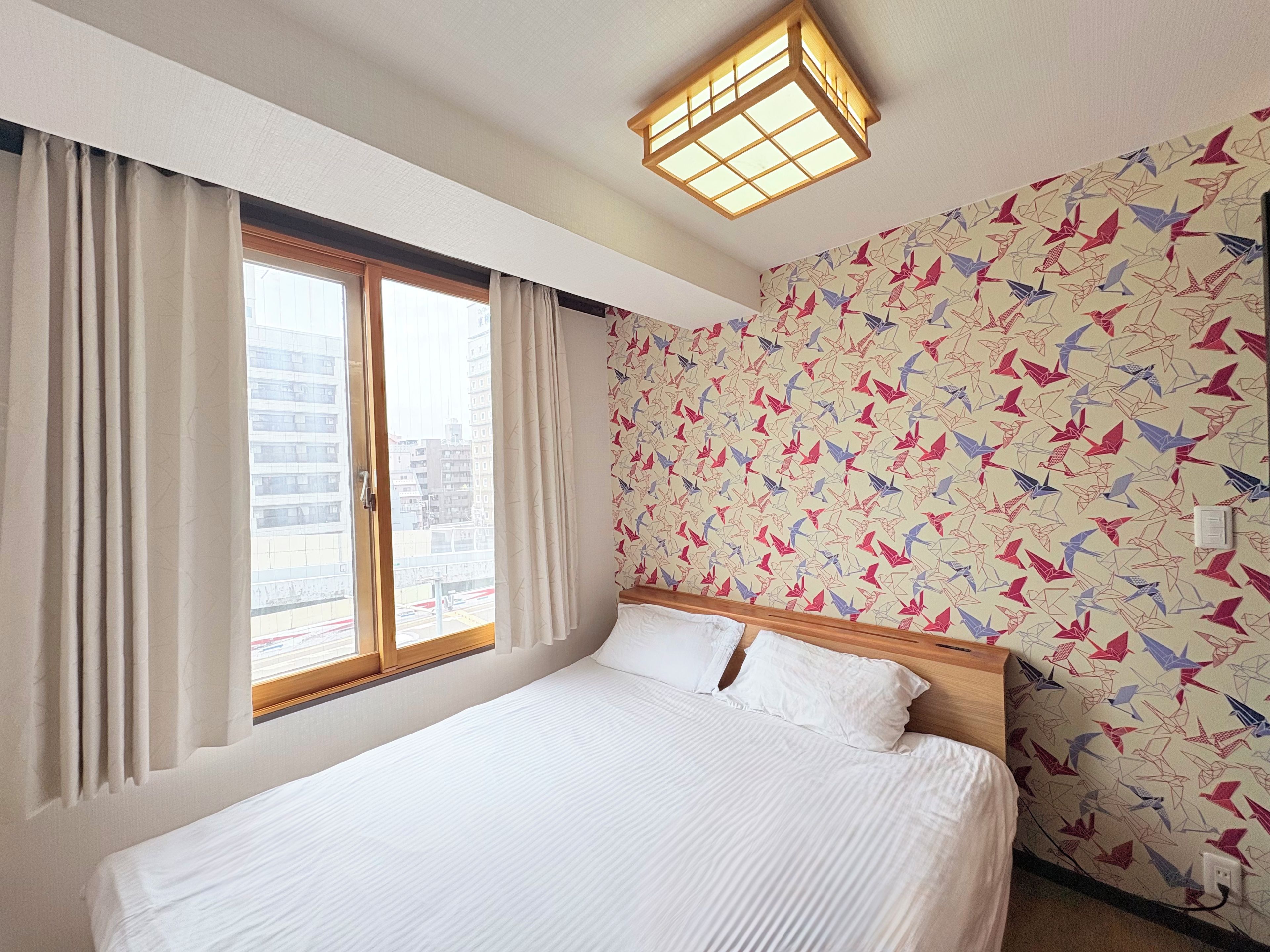 A hotel room with a bed, a window with curtains, a patterned wall, and a ceiling light fixture.