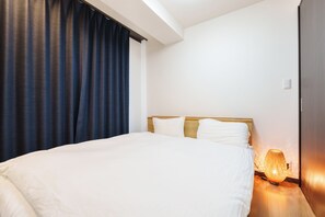 Quarto de hotel com uma cama grande, cabeceira de madeira, mesa de cabeceira com abajur e cortinas escuras.