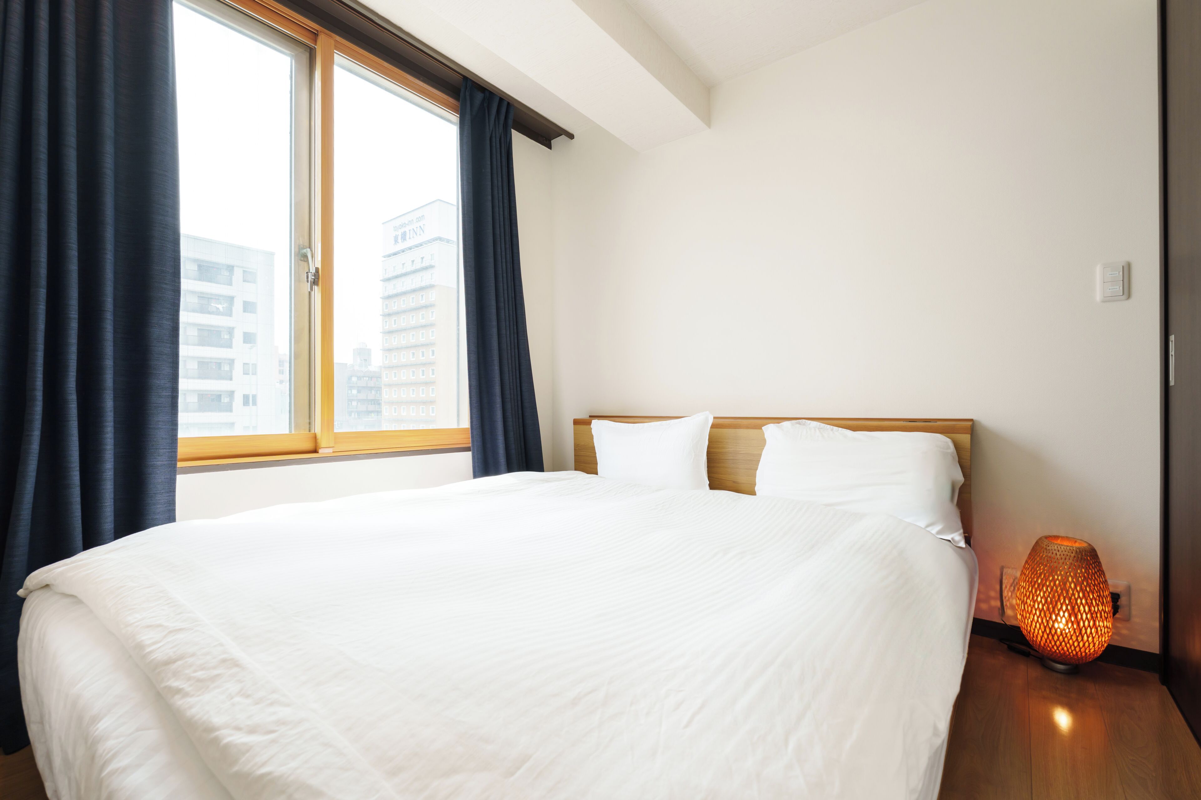 Habitación de hotel con una cama grande, una ventana con cortinas, una lámpara de noche y vistas a edificios.