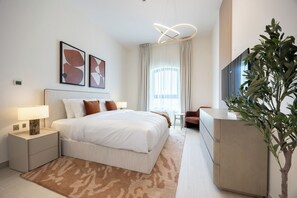 A modern hotel room with a large bed, bedside tables, a desk, a chair, a television, and a plant. - Bloomfields 1BR in Vantage 2-YGC (Abu Dhabi)