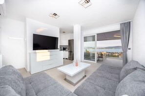 Ein modernes Wohnzimmer mit Flachbildfernseher, grauen Sofas, einem weißen Couchtisch und einem Balkon mit Blick auf die Stadt.