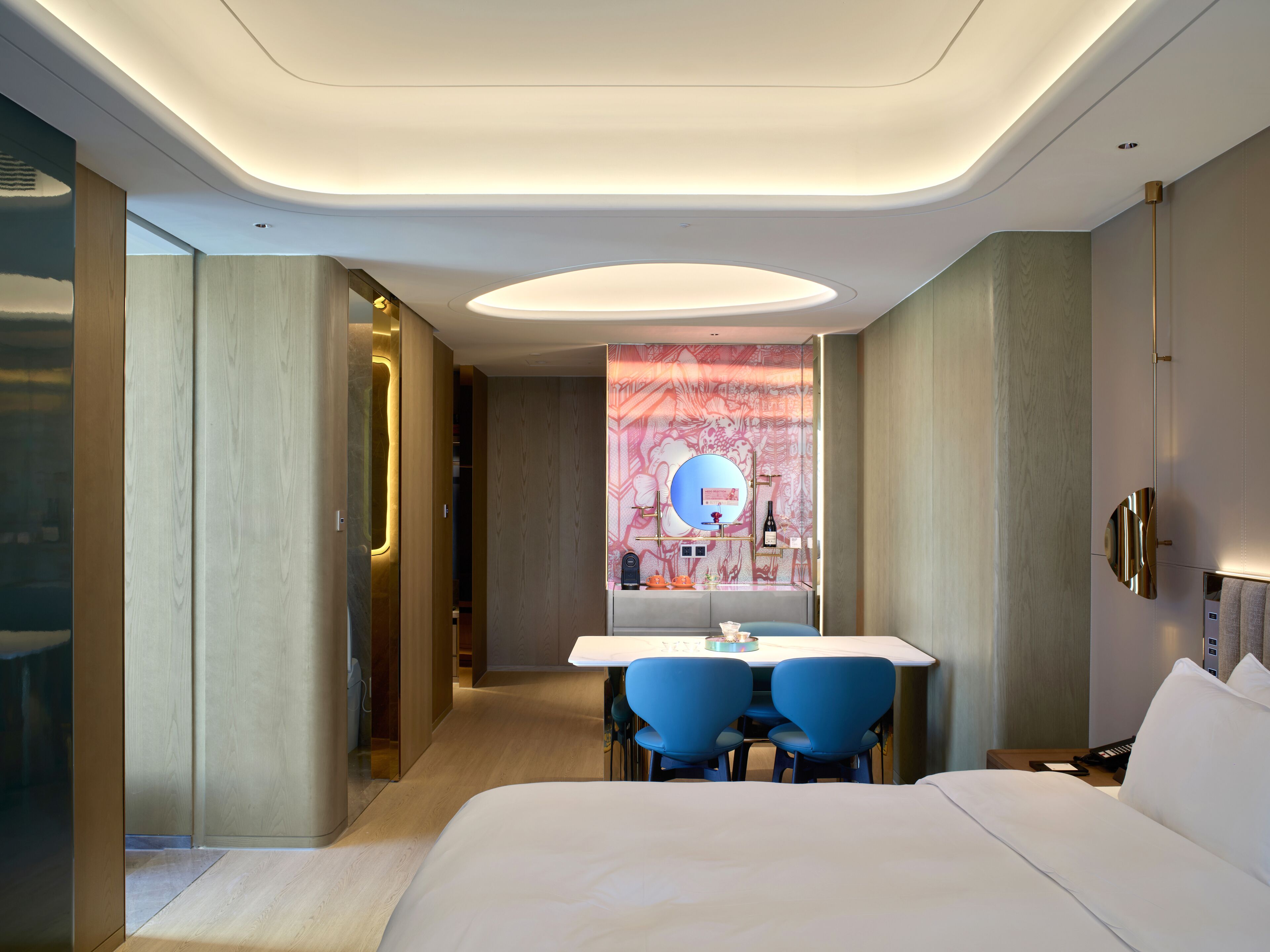 A modern hotel room with a large bed, a desk with two chairs, a vanity with a mirror, and a wall with a decorative pattern.