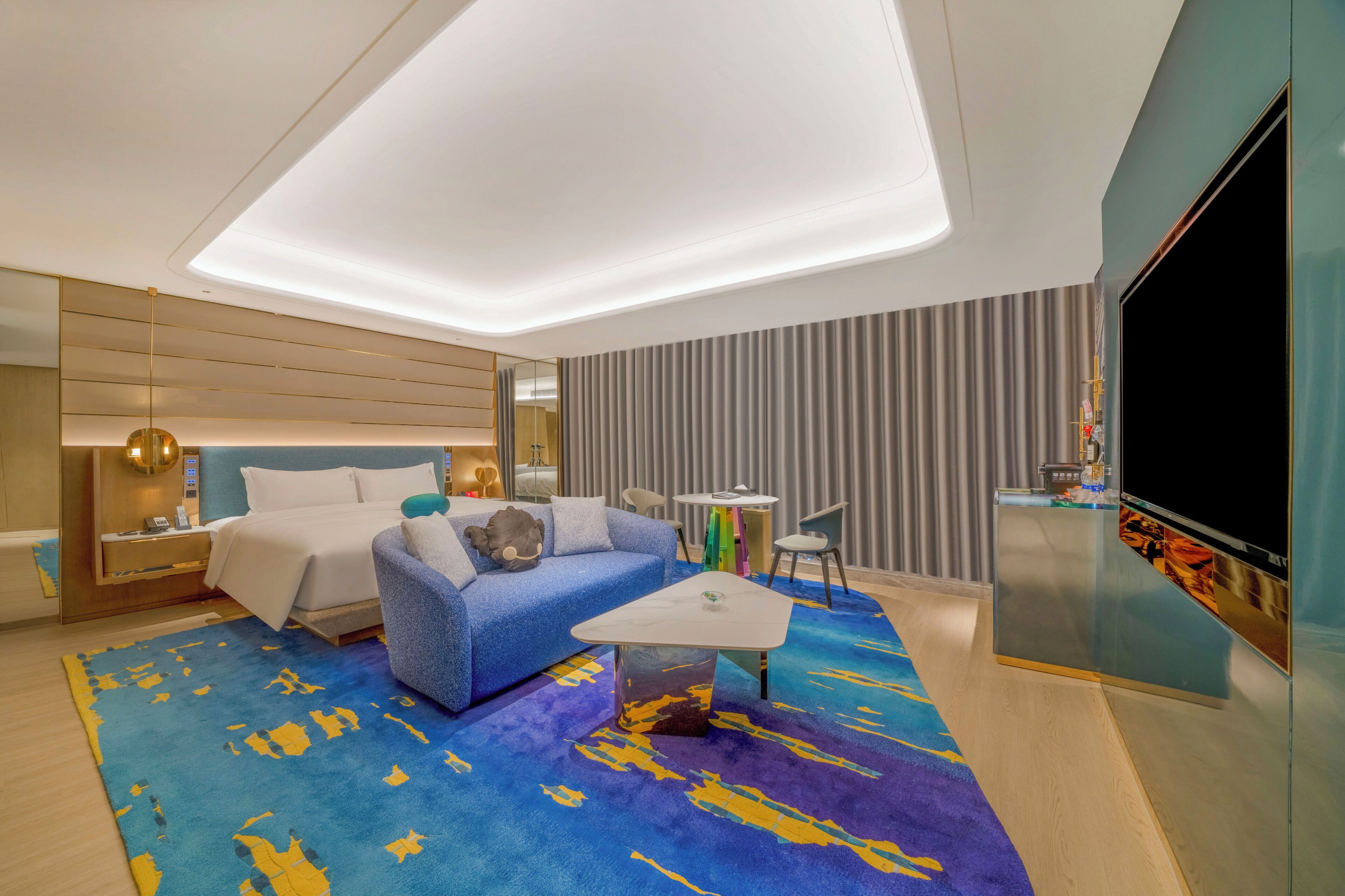 A modern hotel room with a bed, a sofa, a coffee table, a TV, and a minibar.